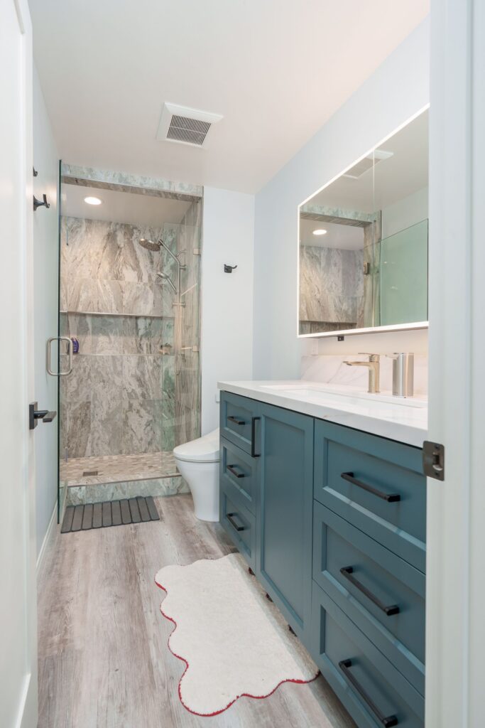 Bathroom remodel with teal vanity and drawers, lighted mirror cabinet, glass-enclosed shower with marble-look tile and long niche