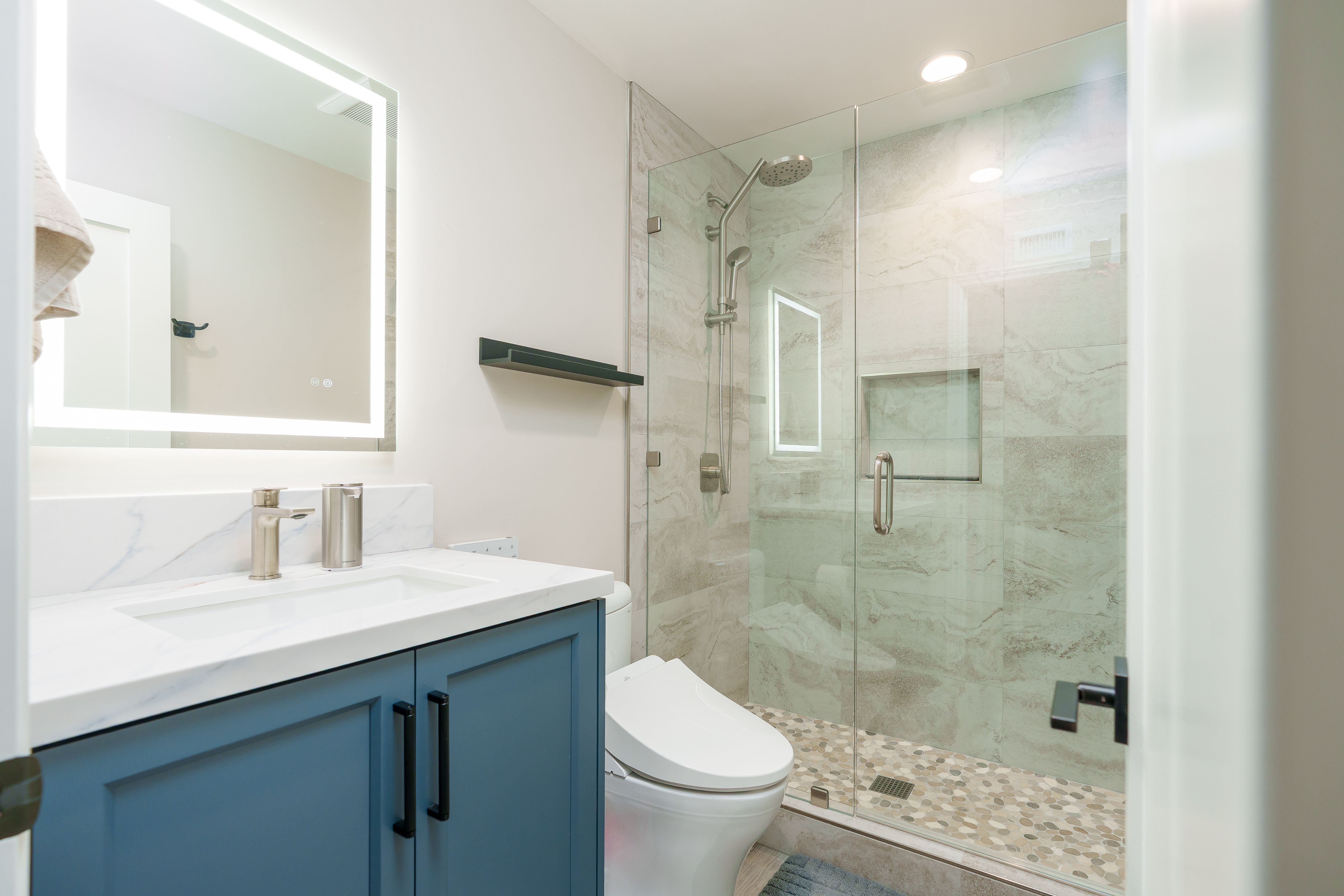 Bathroom with blue shaker vanity, LED mirror, frameless glass shower, stone-look wall tile and pebble shower floor