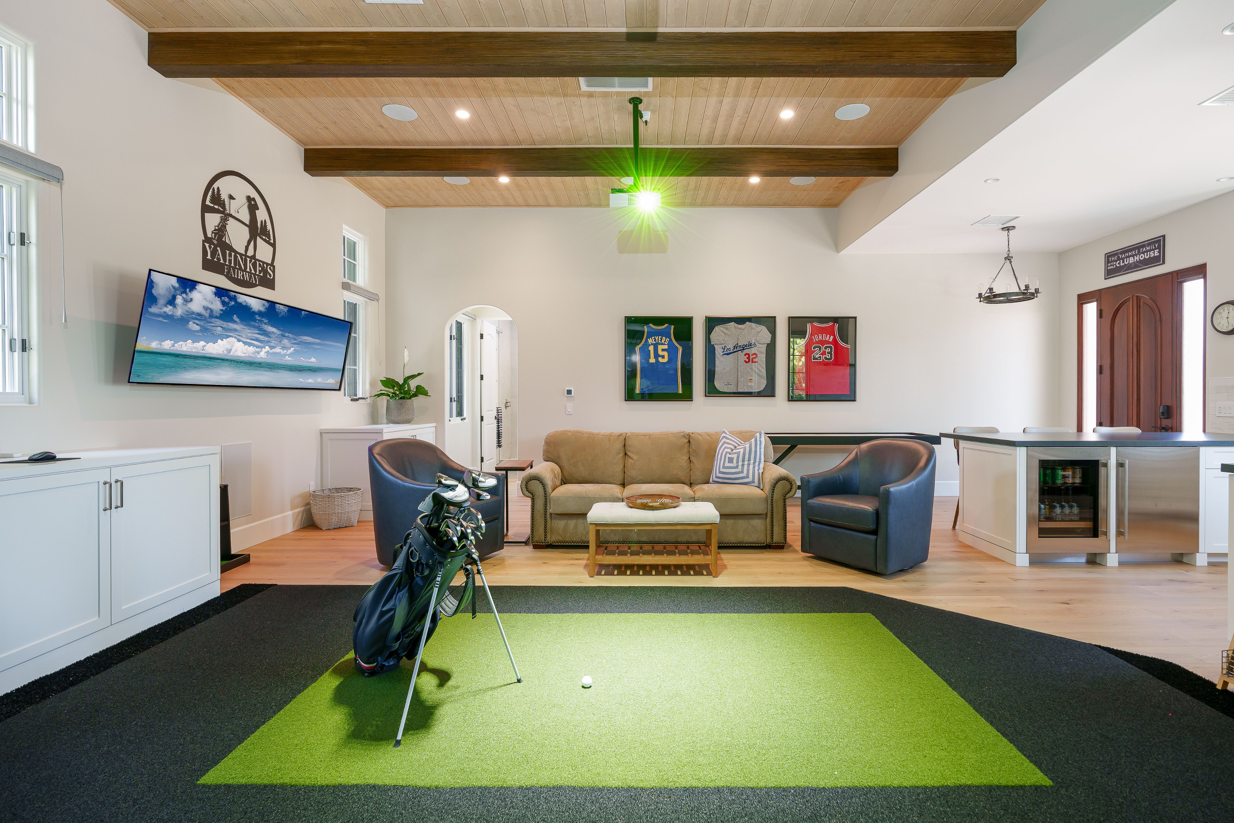 View from simulator turf toward a lounge with leather swivel chairs, framed jerseys, beverage fridge and arched doorway