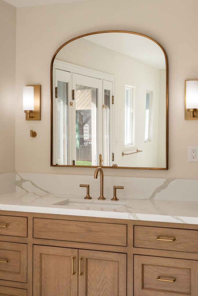 Vanity sink with gold-tone faucet, arched gold-framed mirror, glass-shade sconces and quartz backsplash over wood cabinets
