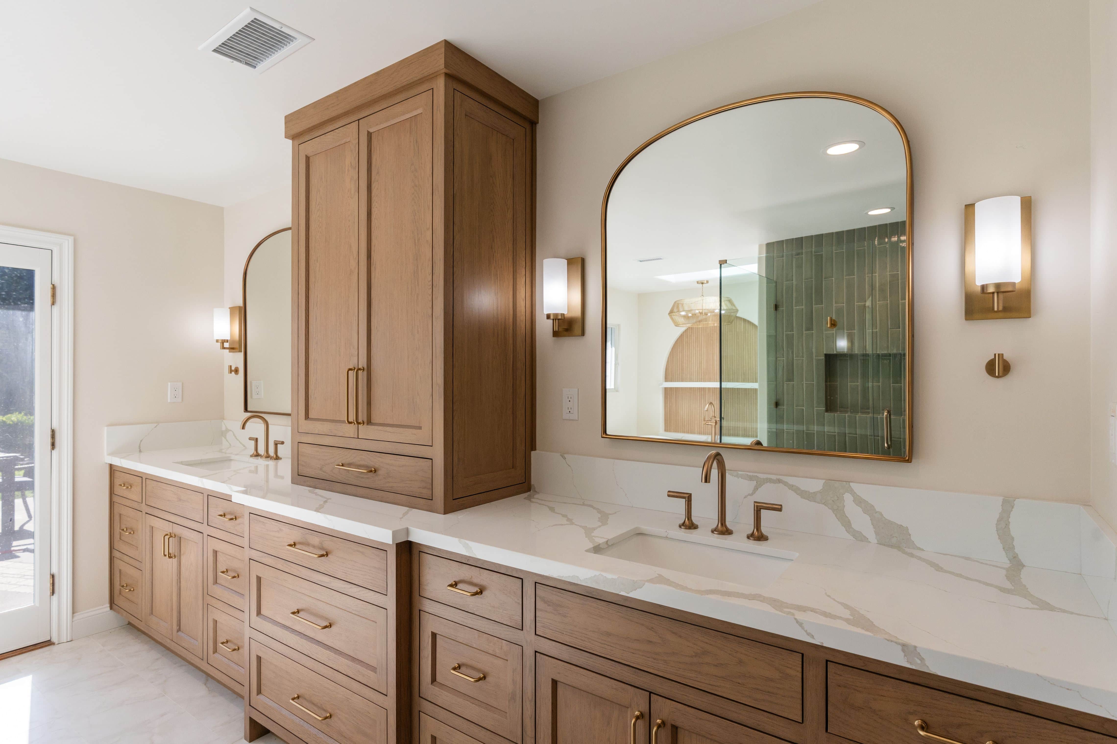 Wood double vanity with quartz top, center tower cabinet, arched gold-framed mirrors and sconces reflecting the shower
