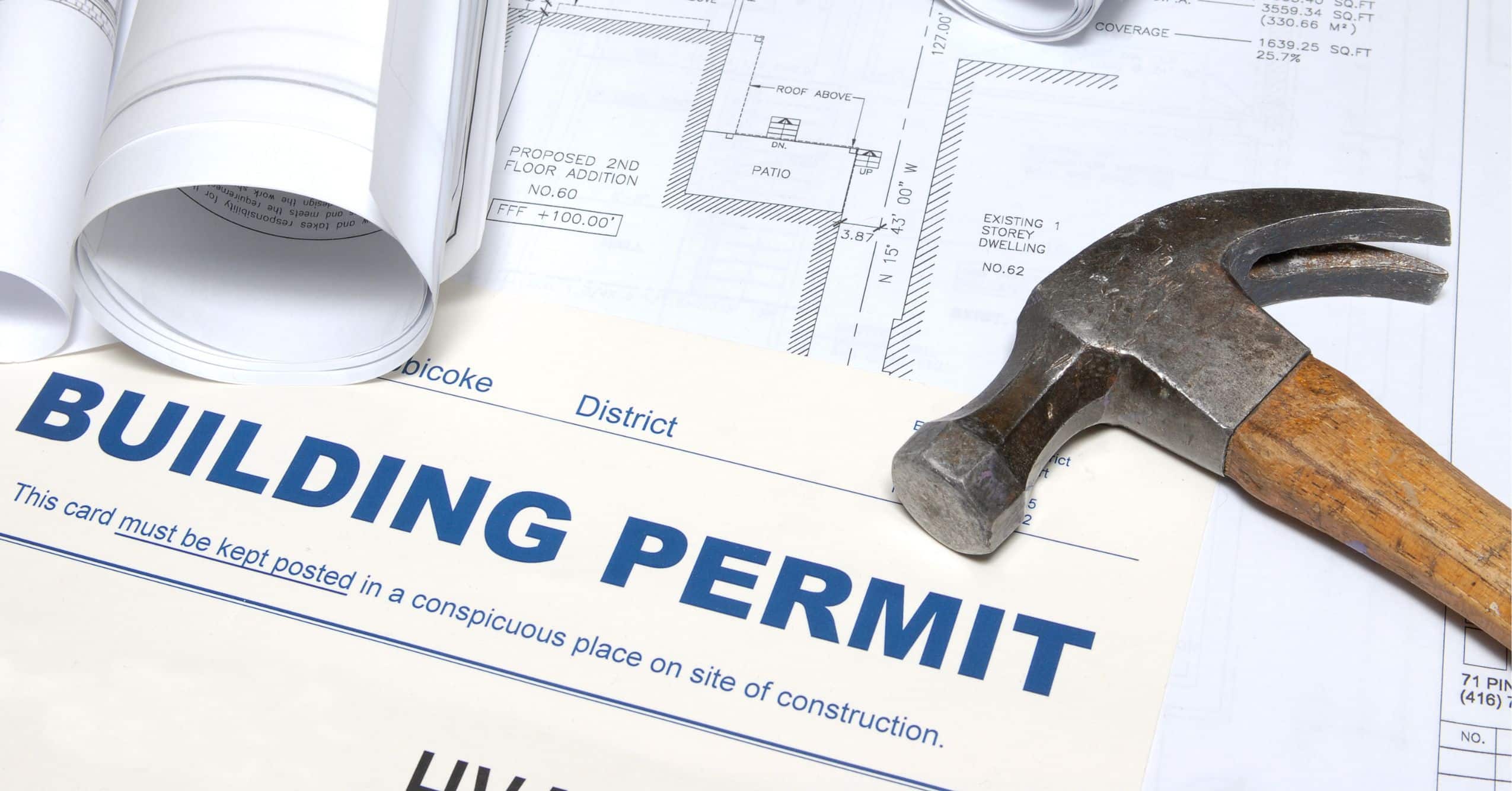 Building permit, architectural plans, rolled drawings, and a hammer arranged on a work surface.