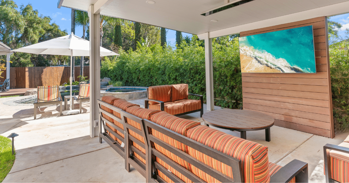 Shaded patio lounge with striped orange cushioned seating, a round table and a wall-mounted TV on a wood slat privacy wall, looking out to a pool and spa.