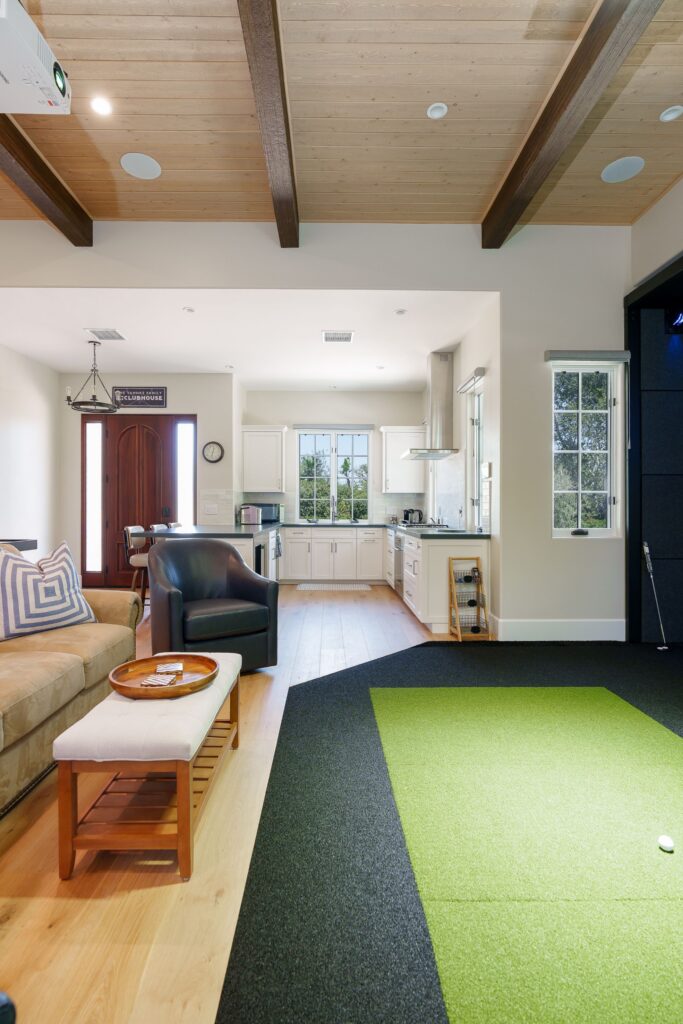 View from the kitchen across wide-plank oak floors to a recessed turf hitting mat, lounge seating and beverage fridge