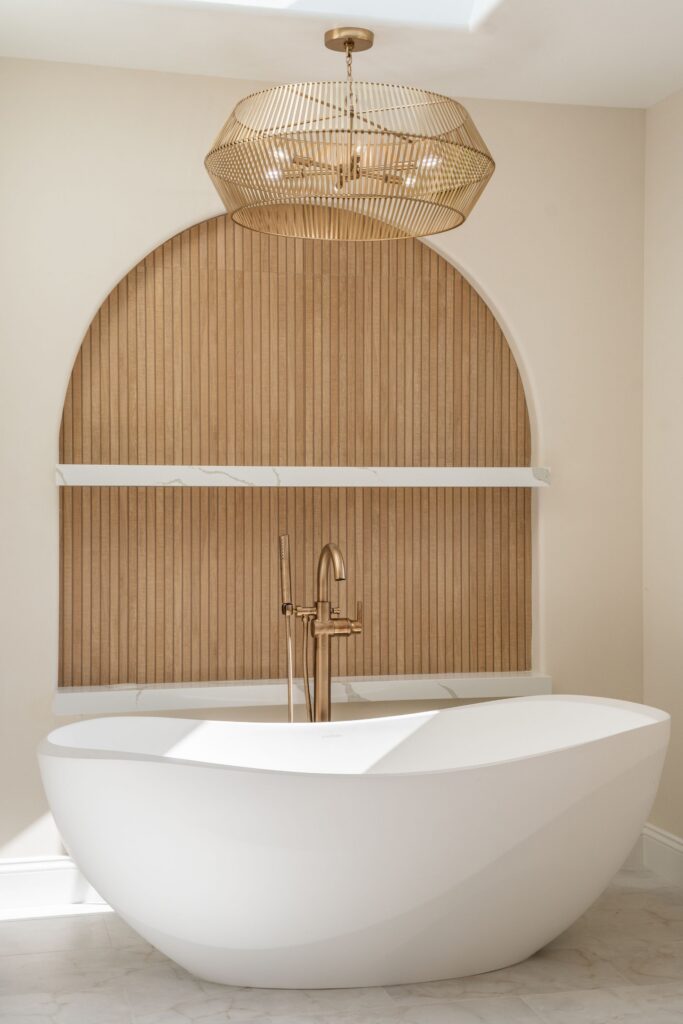 Freestanding soaking tub below a gold cage chandelier in an arched wood-slat alcove with quartz ledges