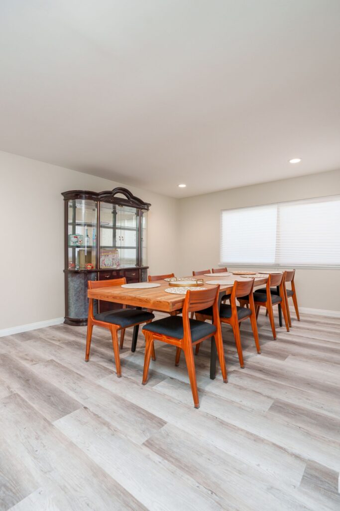 Dining room with long wood table and chairs, china cabinet, wide window with shades, recessed lighting and gray LVP flooring