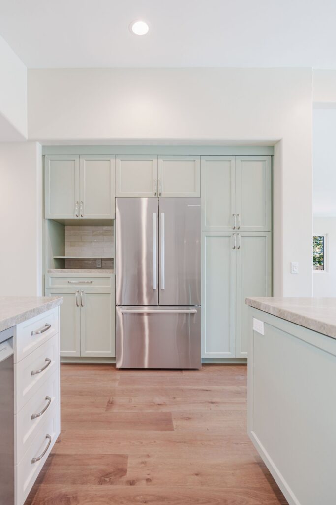 Sage green cabinet wall framing a French-door refrigerator, with tall pantry cabinets and an open appliance nook