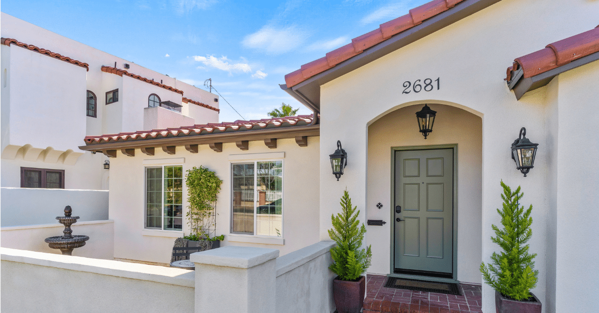 Spanish-style front entry with an arched porch, a sage green door, lantern sconces and a clay tile roof beside a walled courtyard with a tiered fountain.