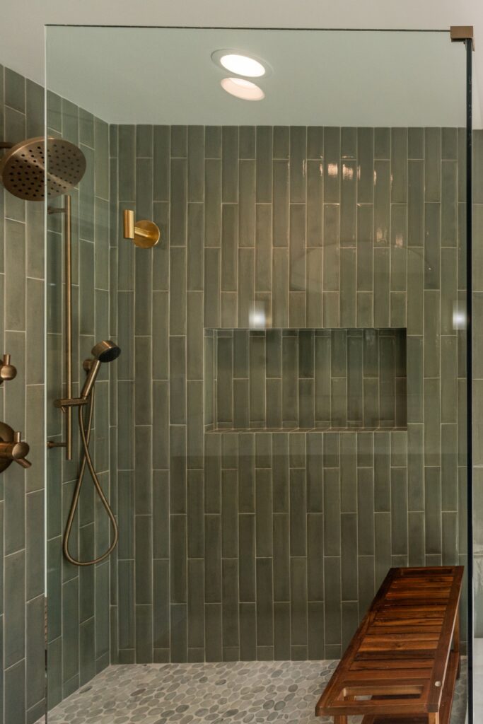 Frameless glass shower with a long tiled niche, green vertical tile walls, slatted wood bench and pebble tile floor