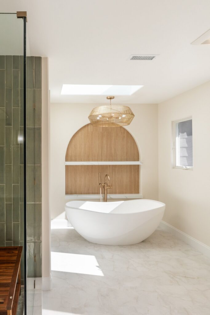 Primary bathroom remodel viewed past the green tile shower to a freestanding tub, skylight and arched wood-slat niche