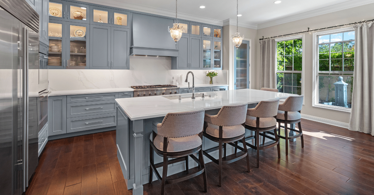 Gray kitchen with large white island, four upholstered stools, glass-front cabinets, and hardwood floors.