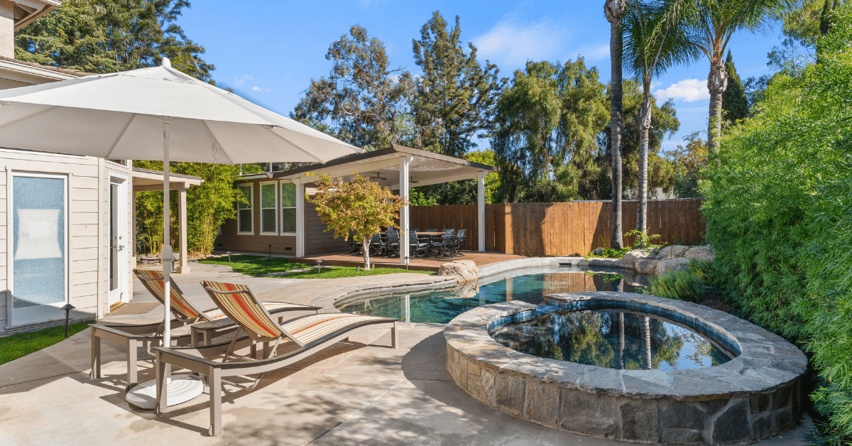 Freeform pool with a raised stone spa, lounge chairs and an umbrella on a concrete deck, with a white patio cover over a dining area in the background.