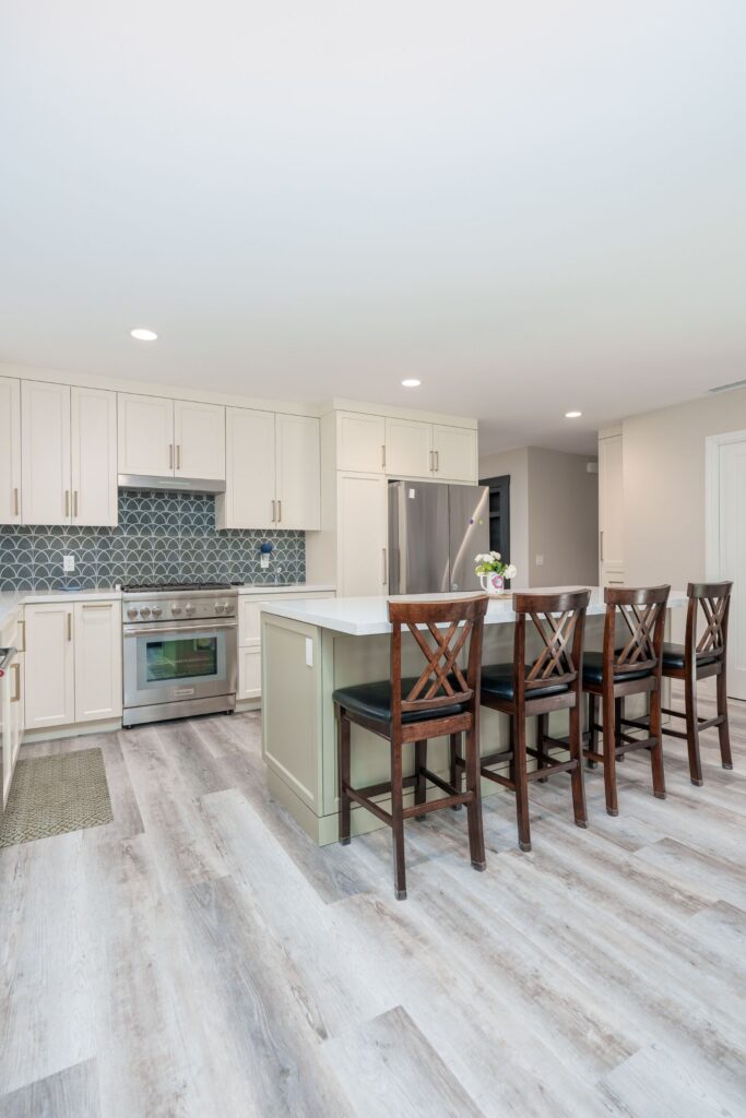 Open kitchen with sage green island, four wood barstools, French-door refrigerator in cabinet surround and recessed lighting