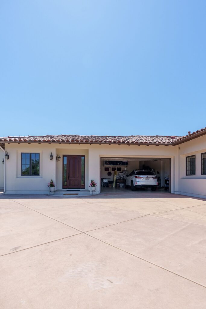 Concrete driveway lined with olive trees and plantings, leading to a stucco ADU with a clay tile roof in San Diego
