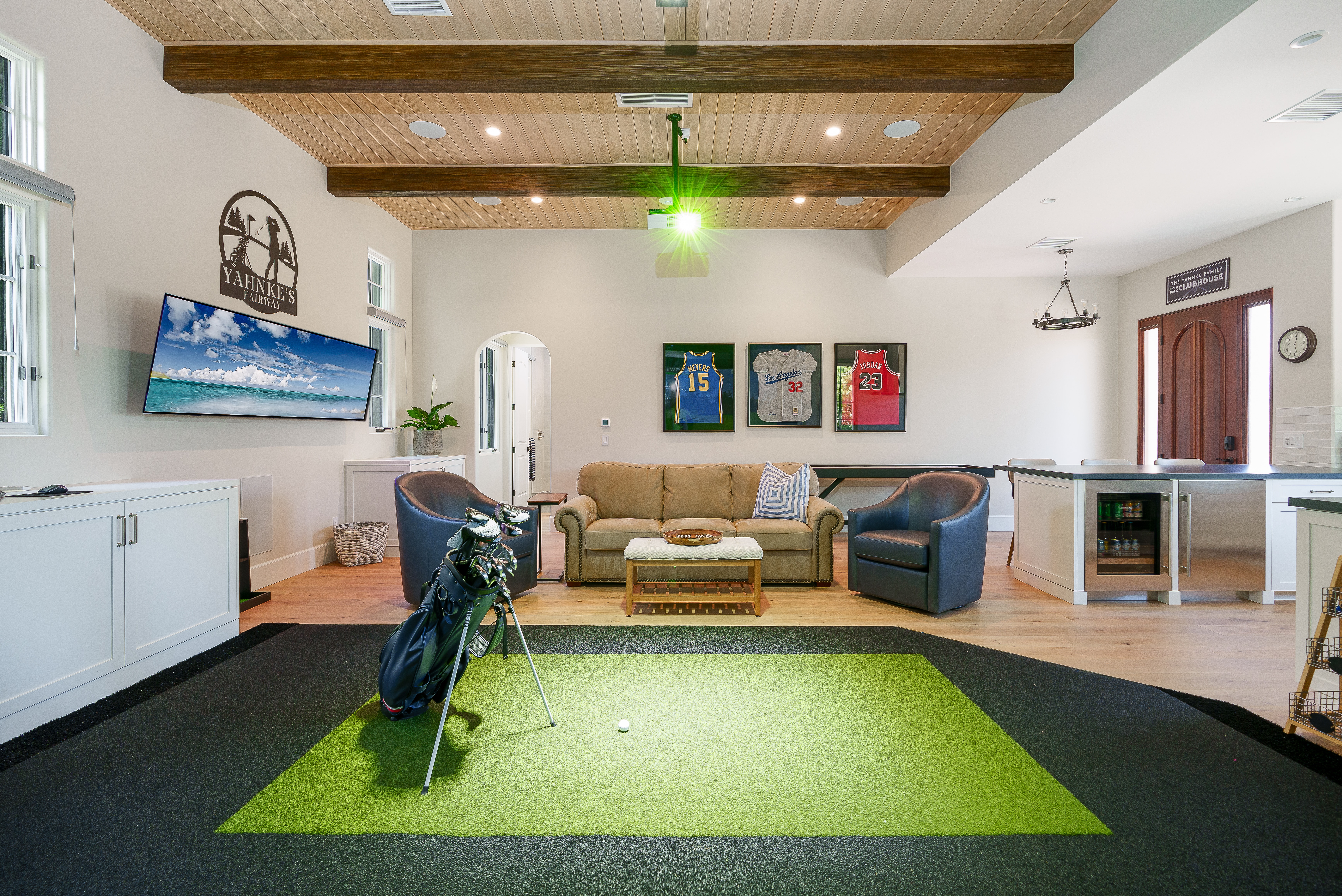 Finished recreation room with indoor golf practice area, seating, wood beam ceiling, television, and kitchenette.