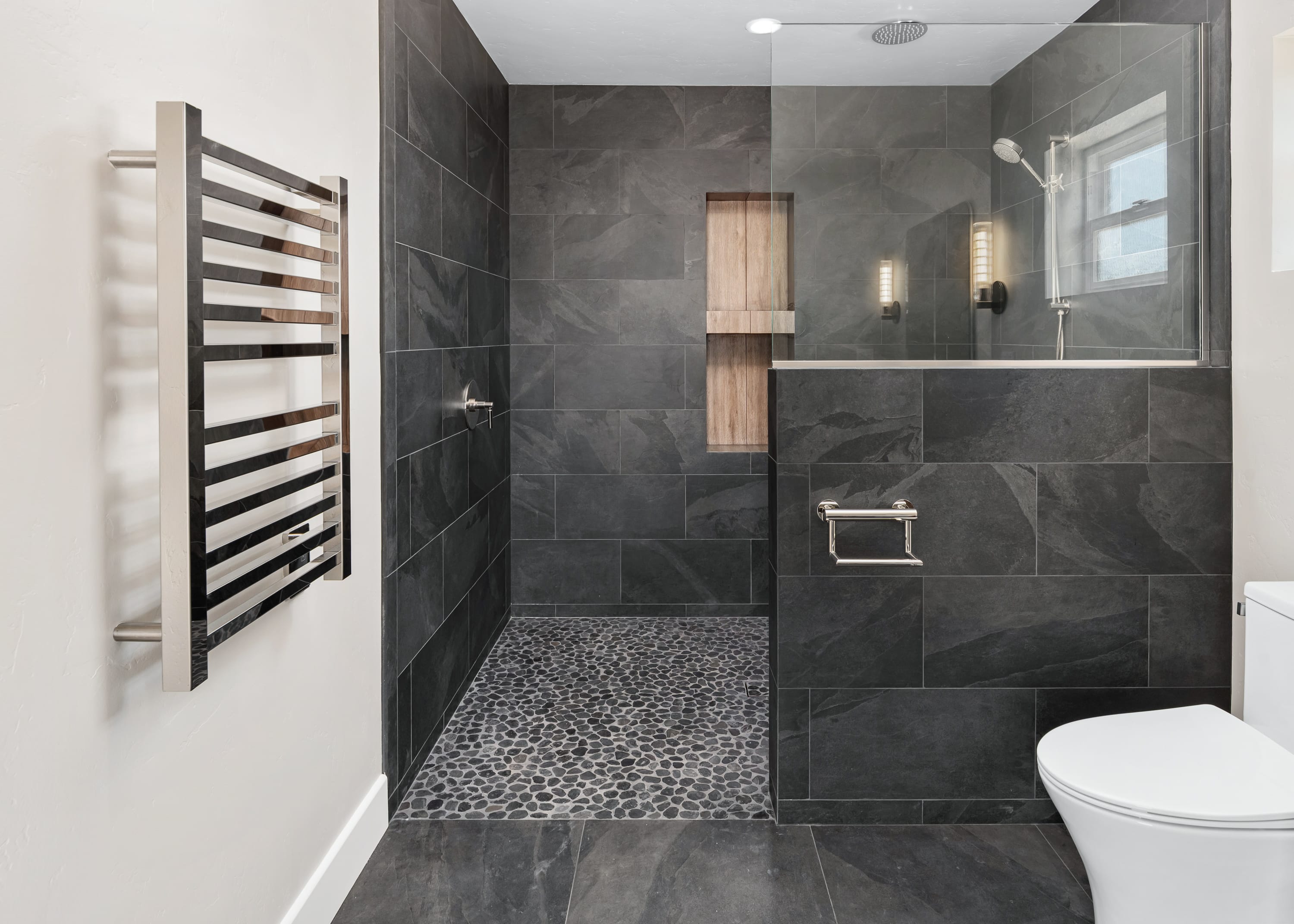 Walk-in shower with large-format charcoal gray tile, pebble stone flooring, glass enclosure, recessed niche, and handheld shower fixture.