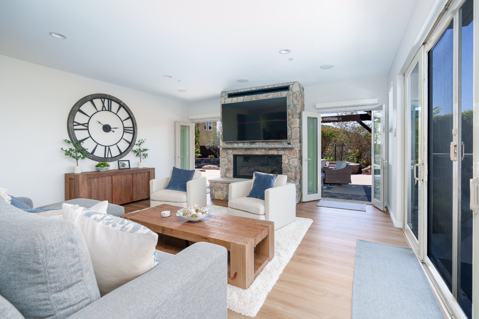 Bright living room with gray sectional, two cream swivel chairs, stone fireplace, open French doors, and sliding glass doors to the patio and view.