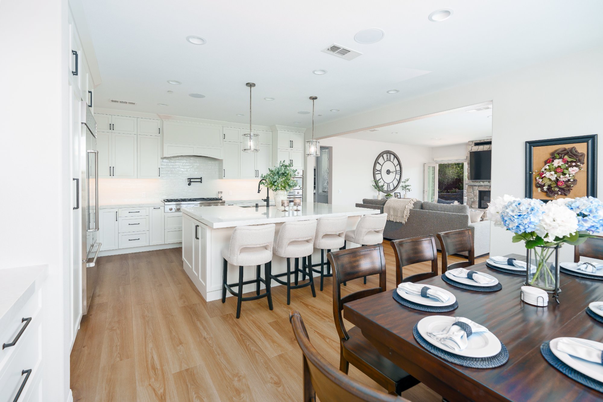 Open first floor with white shaker kitchen, island seating for four, dining table, and living room with stone fireplace beyond.