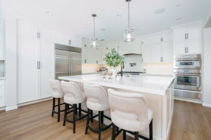 White shaker kitchen with quartz island, four upholstered counter stools, glass pendants, built-in refrigerator, and stacked wall ovens.