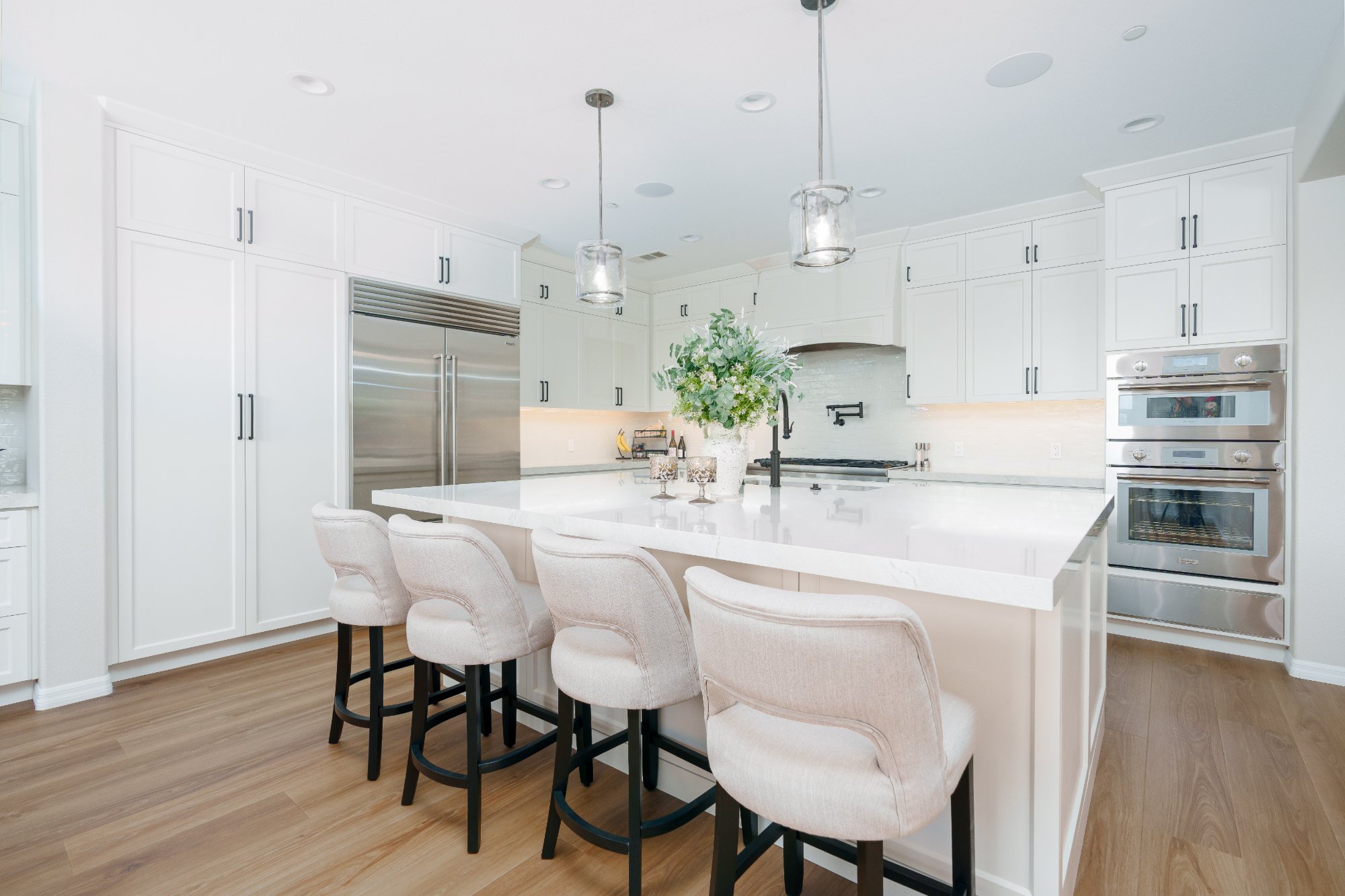 White shaker kitchen with quartz island, four upholstered counter stools, glass pendants, built-in refrigerator, and stacked wall ovens.