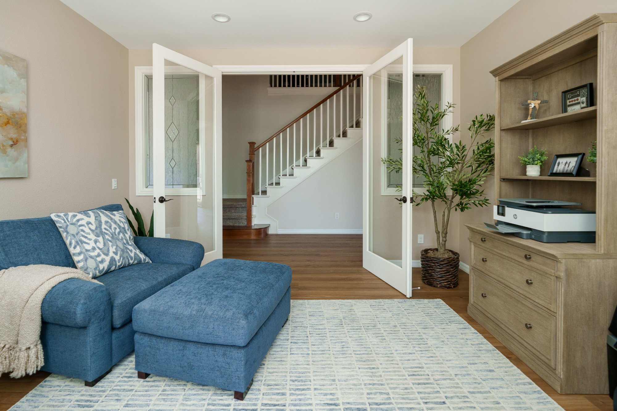 Home office with blue chair and ottoman, area rug, wood storage hutch, and glass French doors opening to the entry and staircase.